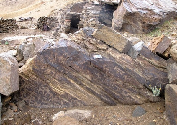 Fig. 12. An ancient naturally-occurring foundation stone pullulating with carvings. In the background are structures of the middle and upper tiers of Rock Art Village. On the boulder are wild yaks and other wild herbivores. There is an anthropomorphic figure visible in the middle of the rock face. Also, note the square with an X in it on the lower right side of the boulder.